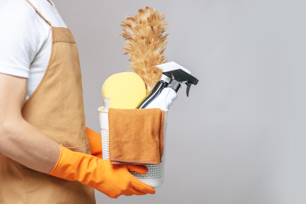side-view-close-up-hand-young-man-apron-rubber-gloves-holding-basket-cleaning-equipment-feather-duster-spray-bottle-sponge-cloth-wiping-basket-min