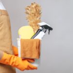 side-view-close-up-hand-young-man-apron-rubber-gloves-holding-basket-cleaning-equipment-feather-duster-spray-bottle-sponge-cloth-wiping-basket-min