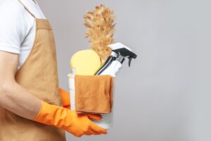 side-view-close-up-hand-young-man-apron-rubber-gloves-holding-basket-cleaning-equipment-feather-duster-spray-bottle-sponge-cloth-wiping-basket-min